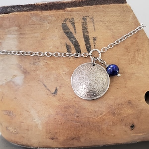 May include: A silver chain necklace with a 1967 British sixpence coin pendant and a dark blue gemstone charm.