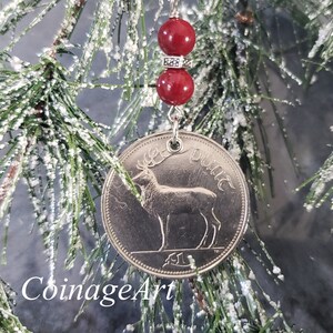 May include: A silver coin ornament featuring a deer design, suspended by a silver chain and red beads. The ornament hangs on a frosted evergreen branch. The text "CoinageArt" is at the bottom.