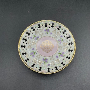 May include: A white porcelain saucer with a delicate, openwork design around the edge. The center features a pink floral pattern with gold accents and purple flowers.