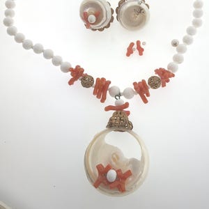 May include: A jewelry set consisting of a necklace and earrings. The necklace features white beads, orange coral branches, gold-toned filigree beads, and a large circular shell pendant with coral and a white bead. Matching shell earrings include coral.
