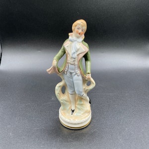 May include: A porcelain figurine of a man in a green and pink suit with a white cravat. He is standing on a base with a gold trim.