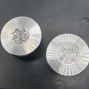 May include: Two circular, brushed metal objects with intricate, raised floral designs in the center. The objects are silver in color and appear to be decorative or functional components.