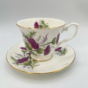 May include: White teacup and saucer set with a gold rim. The cup and saucer are decorated with purple thistle flowers and green leaves. The teacup has a curved handle.
