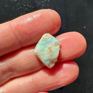 K&ouml;nnte beinhalten: Ein kleiner, grob geschnittener Opal-Edelstein mit einer Mischung aus Farben, darunter Blau, Gr&uuml;n und Wei&szlig;. Der Opal wird zwischen zwei Fingern gehalten und zeigt seine nat&uuml;rliche, unregelm&auml;&szlig;ige Form und sein schillerndes Farbspiel.