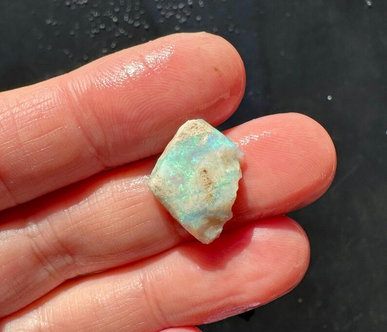 K&ouml;nnte beinhalten: Ein kleiner, grob geschnittener Opal-Edelstein mit einer Mischung aus Farben, darunter Blau, Gr&uuml;n und Wei&szlig;. Der Opal wird zwischen zwei Fingern gehalten und zeigt seine nat&uuml;rliche, unregelm&auml;&szlig;ige Form und sein schillerndes Farbspiel.