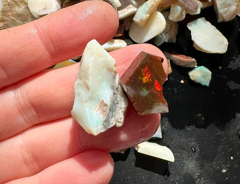 K&ouml;nnte beinhalten: Nahaufnahme von zwei rohen Opal-Edelsteinen. Einer ist hellcremefarben mit Andeutungen von Blau und Gr&uuml;n, der andere ist dunkelbraun mit leuchtenden roten, orangen und gr&uuml;nen Blitzen. Die Edelsteine werden in einer Hand gehalten.