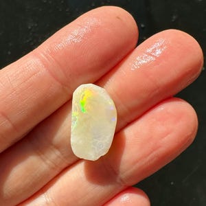 K&ouml;nnte beinhalten: Ein kleiner, grob geschliffener Opal-Edelstein mit cremewei&szlig;er Grundfarbe. Der Opal zeigt schillernde Farben, darunter Gr&uuml;n, Gelb und Lila. Der Stein wird zwischen zwei Fingern gehalten, was seine nat&uuml;rliche Textur und sein einzigartiges Farbspiel zeigt.