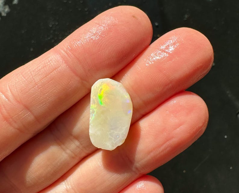 K&ouml;nnte beinhalten: Ein kleiner, grob geschliffener Opal-Edelstein mit cremewei&szlig;er Grundfarbe. Der Opal zeigt schillernde Farben, darunter Gr&uuml;n, Gelb und Lila. Der Stein wird zwischen zwei Fingern gehalten, was seine nat&uuml;rliche Textur und sein einzigartiges Farbspiel zeigt.