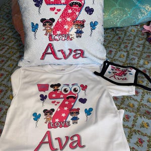 May include: A white sequin pillow, a white t-shirt, and a white face mask, each featuring a graphic of a large pink number seven, cartoon dolls, blue and purple balloons, and the name Ava in pink lettering with the word LOVE printed below.