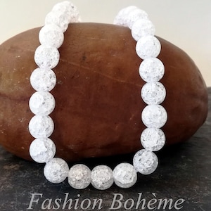 Peut inclure: Un bracelet en perles de cristal blanc avec une surface texturée. Le bracelet est composé de perles rondes et est posé sur une roche brune.