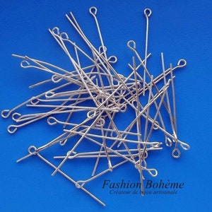 Op de afbeelding: Een stapel zilverkleurige kopspelden met lussen aan het ene uiteinde. De spelden liggen verspreid op een blauwe achtergrond.