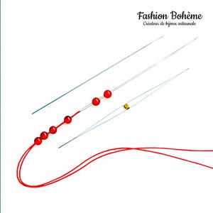 May include: Three beading needles with red beads strung on one of the needles. The needles are silver and the beads are red. A red thread is looped around the needles.