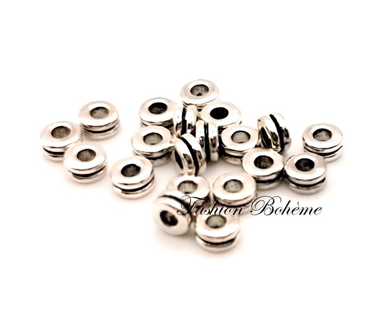 May include: A collection of silver-colored spacer beads. Each bead is cylindrical with a central hole and two grooved bands. The text "Fashion Boh&egrave;me" is visible in the lower right corner.