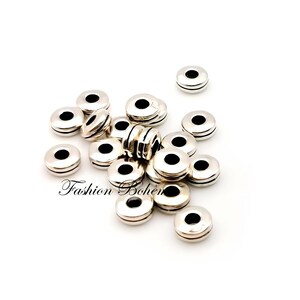 May include: A collection of silver-colored, round beads with a central black hole. The beads have a flat, disc-like shape with a grooved edge. The words "Fashion Bohem" are printed in cursive on the bottom left of the image.