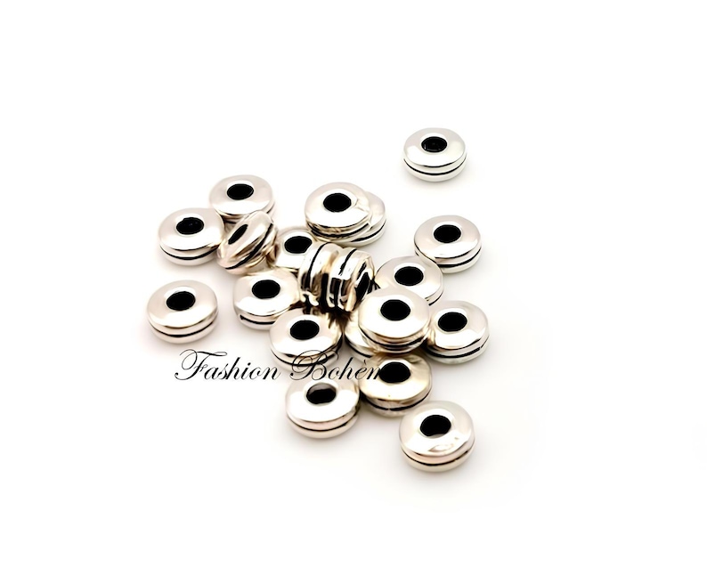 May include: A collection of silver-colored, round beads with a central black hole. The beads have a flat, disc-like shape with a grooved edge. The words "Fashion Bohem" are printed in cursive on the bottom left of the image.