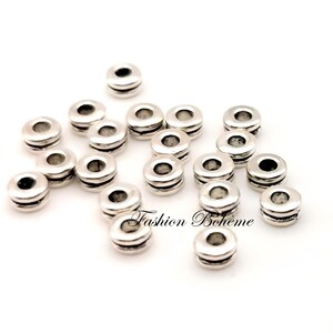 May include: Silver-colored spacer beads with a cylindrical shape and a central hole. The beads have a layered design with two horizontal grooves. The image includes the text "Fashion Boh&egrave;me".