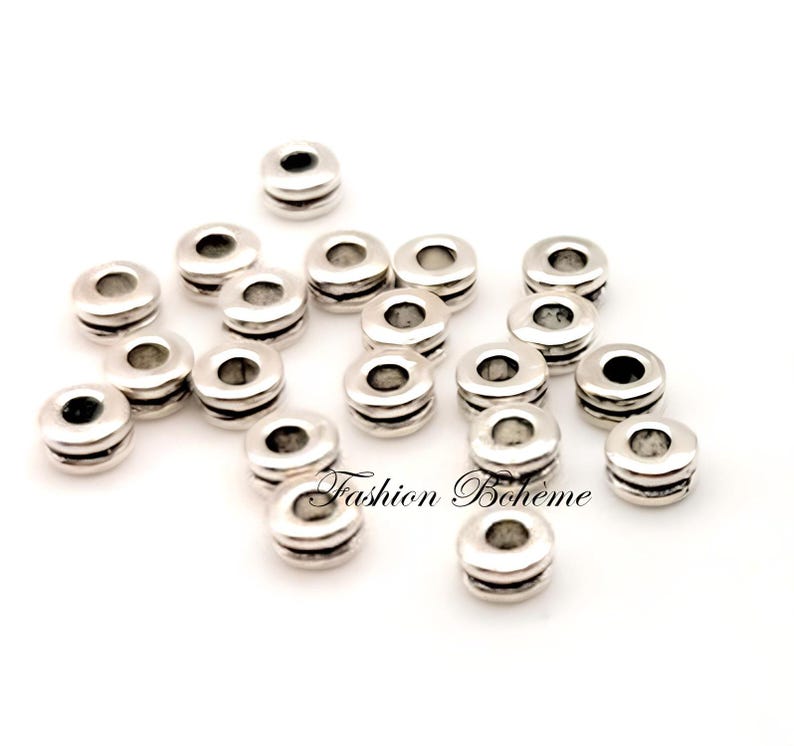 May include: Silver-colored spacer beads with a cylindrical shape and a central hole. The beads have a layered design with two horizontal grooves. The image includes the text "Fashion Boh&egrave;me".