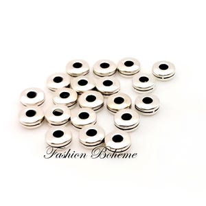 Peut inclure: Plusieurs perles rondes argentées avec un trou central noir et un design rainuré sont disposées sur une surface blanche. Le texte "Fashion Boheme" est imprimé en cursive. Idéal pour la création de bijoux.