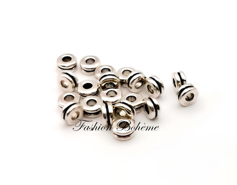 May include: A collection of silver-colored metal beads with a cylindrical shape and a central hole. The beads have a grooved design around the middle. The text "Fashion Boh&egrave;me" is visible on the bottom of the image.