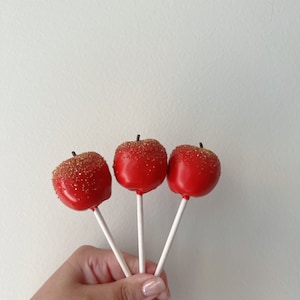 May include: Three red apple-shaped cake pops with gold sprinkles on white sticks held in a hand.
