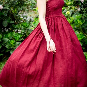 May include: A burgundy-colored dress with a fitted bodice and a flowing skirt. The dress features a sweetheart neckline, wide straps, and a lace-up detail on the side. The dress is made of a soft, flowing fabric, perfect for a special occasion.