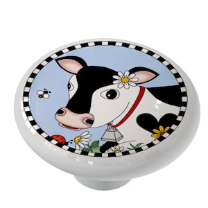 May include: A white ceramic knob with a black and white cow face design. The cow has a red collar and a bell. The background is blue with white daisies and a ladybug.