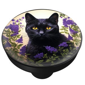 May include: A black cat with yellow eyes is the central image on a round, black phone grip. The cat is surrounded by purple flowers and green leaves against a cream background. The phone grip is a practical and decorative accessory.