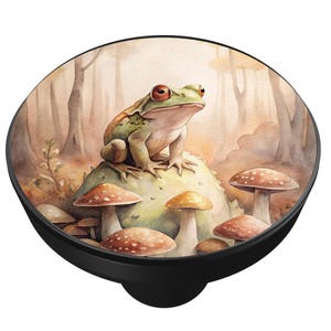 May include: A round, black PopSocket featuring a watercolor illustration of a green frog with red eyes perched on a mossy rock surrounded by mushrooms. The background depicts a forest scene with tall trees and a soft, muted color palette.