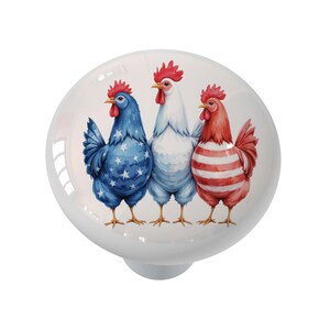 May include: A white ceramic cabinet knob featuring three stylized roosters. The rooster on the left is blue with white stars, the center rooster is white, and the right rooster is red with white stripes. The knob has a glossy finish.