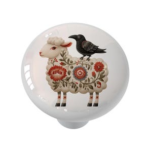 May include: White ceramic cabinet knob featuring a sheep with floral patterns and a black crow. The sheep has red, blue, and cream-colored floral designs. The knob is round and has a metal base.