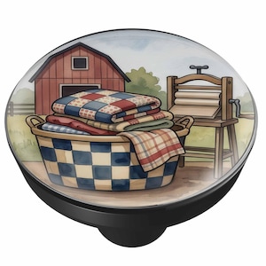 May include: A round cabinet knob featuring a printed illustration of a blue and white checkered laundry basket filled with folded quilts. In the background, a red barn and a wooden wringer washing machine are visible under a light sky.