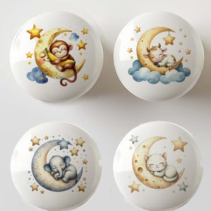 May include: Four white ceramic cabinet knobs with whimsical illustrations. Each knob features a different animal: a monkey with a banana, a cow on clouds, an elephant, and a cat, all sleeping on crescent moons surrounded by stars.