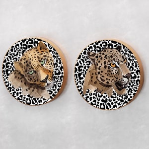 May include: Two round coasters with a black and white leopard print background. Each coaster features a watercolor painting of a leopard's face with green eyes and orange spots.