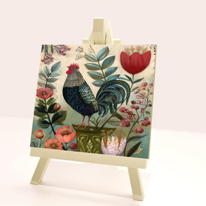 May include: A decorative painting on a small easel features a rooster with blue and black plumage, standing amidst colorful flowers and foliage. The artwork includes a large red tulip and various pink blossoms, set against a cream-colored background.