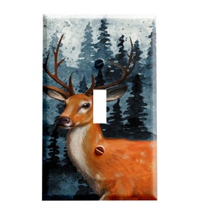 May include: A decorative light switch cover featuring a watercolor painting of a deer with large antlers. The deer is a warm orange color with a white chest, set against a backdrop of dark blue evergreen trees.