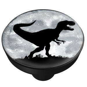 May include: A black dinosaur silhouette against a gray, moonlit background, set on a black, round knob. The dinosaur is a Tyrannosaurus Rex, with its mouth open and tail curved. The knob is likely for a cabinet or drawer.