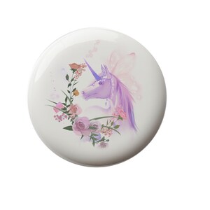 May include: A white, round decorative item featuring a purple unicorn with a flowing mane and a horn. The unicorn is surrounded by a floral wreath of pink and peach roses and greenery. The design has a whimsical, fairytale theme.