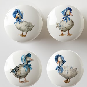 May include: Four white ceramic cabinet knobs with a goose illustration. Each knob features a different goose wearing a blue bonnet with white polka dots and a matching bow. The geese have orange beaks and feet.