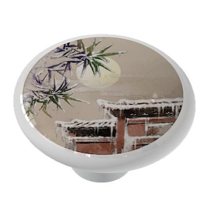 May include: A ceramic knob with a watercolor painting of a snowy scene with a building and a tree. The building has a red roof and the tree has green leaves. The background is a light brown color.