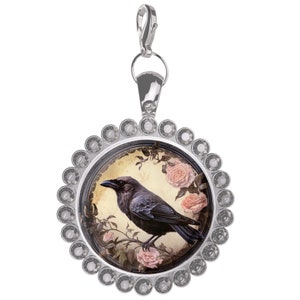May include: A silver charm with a round, clear crystal-encrusted frame. Inside the frame is a vintage-style illustration of a black raven perched on a branch with pink roses.