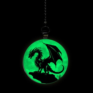 May include: A black silhouette of a dragon with wings spread wide, perched on a rock, against a glowing green moon background. The image is on a round pendant with a silver chain.