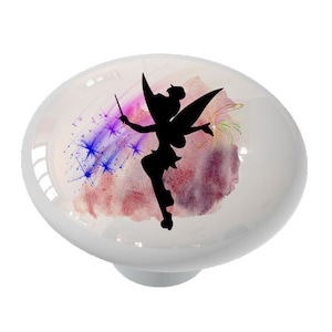 May include: White ceramic cabinet knob featuring a black silhouette of a fairy with a wand, set against a watercolor background of pink, purple, and blue. The knob has a glossy finish and a simple, rounded shape.