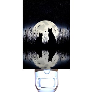 May include: A nightlight featuring a black and white image of two cats silhouetted against a full moon and starry sky reflected in a body of water.