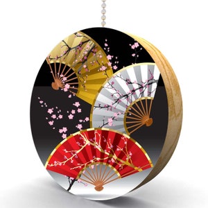 May include: A round wooden ornament with a black background and three Japanese fans with cherry blossom designs. The fans are red, gold, and white.
