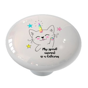 May include: White ceramic knob with a cartoon cat with a unicorn horn. The cat is smiling and has a paw print on its paw. The text "My spirit animal is a Caticorn" is below the cat.