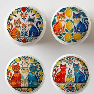 May include: Four round decorative cabinet knobs with colorful cat designs. Each knob features a pair of stylized cats in orange, red, blue, and white, surrounded by floral and lemon motifs on a white background. The knobs are likely made of ceramic.