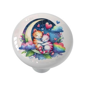 May include: White ceramic cabinet knob featuring a watercolor illustration of two hugging cats on a crescent moon. The design includes hearts, clouds, and a starry night sky, with a rainbow accent. A decorative accent for furniture.