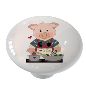 May include: A ceramic knob with a pink pig wearing a red apron and holding a spatula and a bottle of seasoning. The pig is cooking in a kitchen with a pot and a pan on the stove. The text on the apron says "I love to cook".