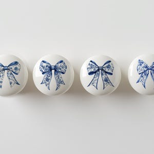 May include: Four white ceramic cabinet knobs with a blue floral bow design. The bows have intricate details and are centered on the knobs. The knobs are arranged in a row against a white background.