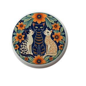 May include: A round decorative ceramic tile featuring three stylized cats. The cats are set against a backdrop of orange flowers and green leaves. One cat is black and white, one is black and orange, and the other is orange and white.